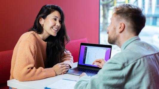 Woman and man sit across from each other at desk, laptop in between them. Man is pointing at something on laptop, woman smiling.